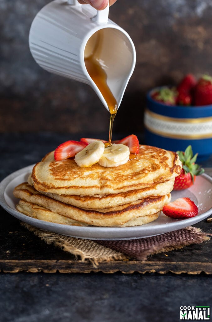 maple syrup being poured over a stack of pancakes topped with strawberries and banana