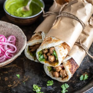 three wraps rolled in parchment paper and tied with thread stacked together with a bowl of chutney and pickled onions on the side