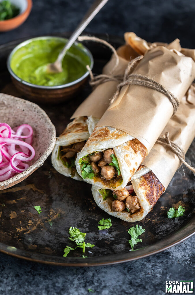 three wraps rolled in parchment paper and tied with thread stacked together with a bowl of chutney and pickled onions on the side
