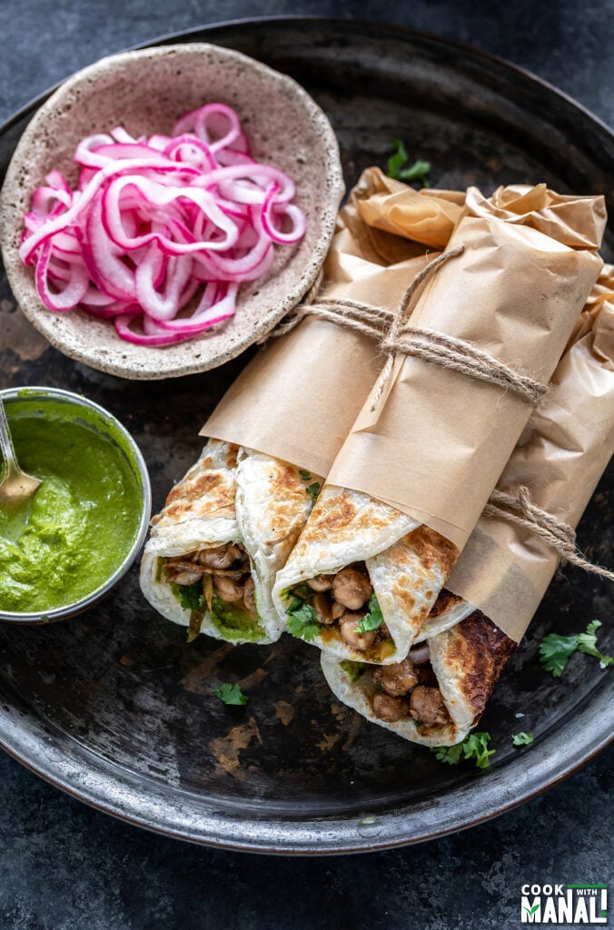 three wraps rolled in parchment paper and tied with thread stacked together with a bowl of chutney and pickled onions on the side