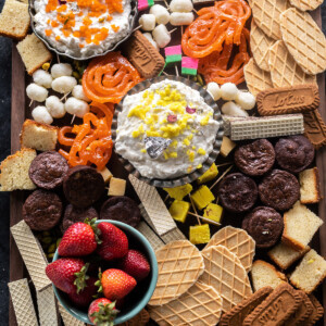 a dessert board filled with dips, mithai, fruits, cake, brownies