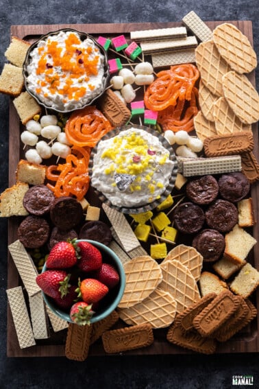 a dessert board filled with dips, mithai, fruits, cake, brownies