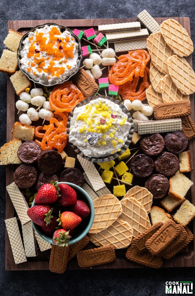 a dessert board filled with dips, mithai, fruits, cake, brownies