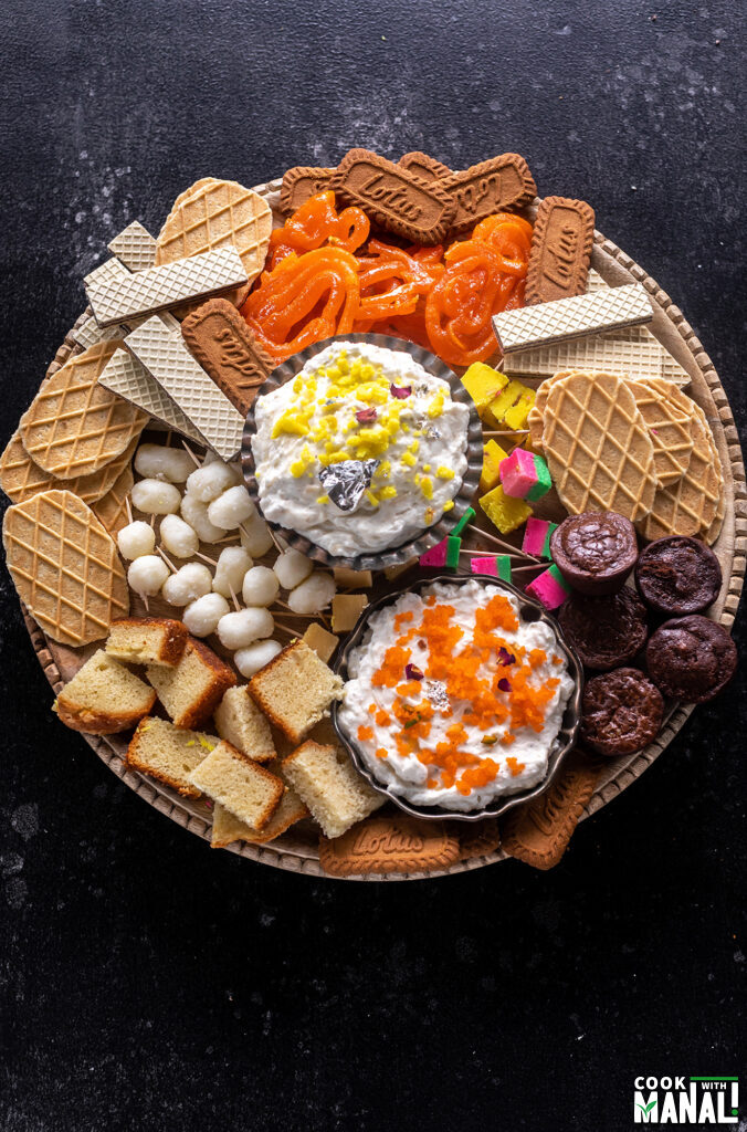 a dessert board filled with dips, mithai, fruits, cake, brownies