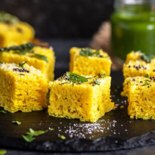 khaman dhokla arranged on a black board with bottle of chutney in the backdrop