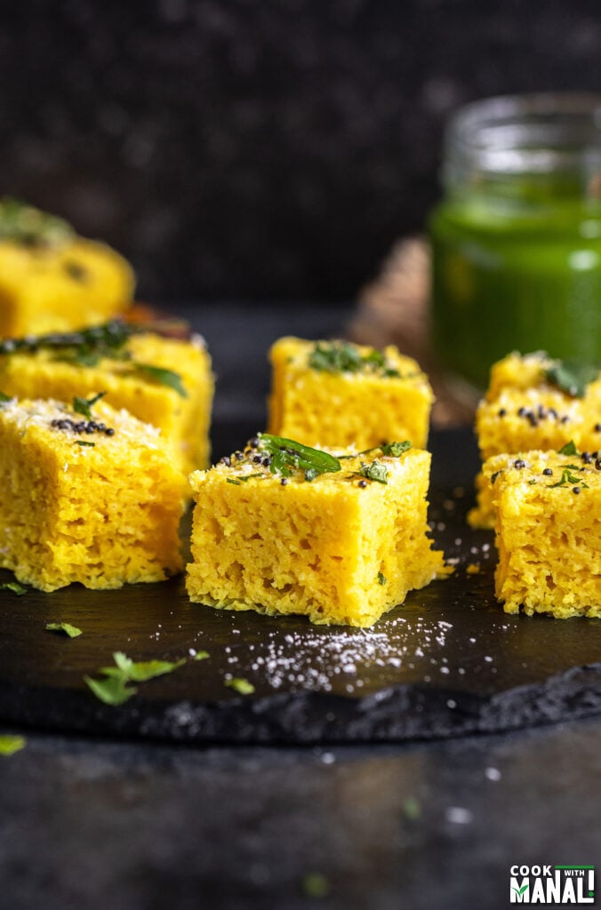 khaman dhokla arranged on a black board with bottle of chutney in the backdrop