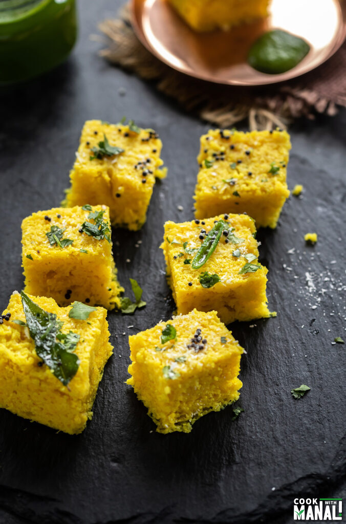 cut dhokla pieces arranged on a black board topped with curry leaves, cilantro and coconut