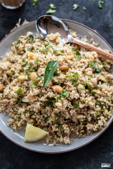 Quinoa upma served in a plate topped with curry leaves, lemon wedge and a copper spoon placed on the side