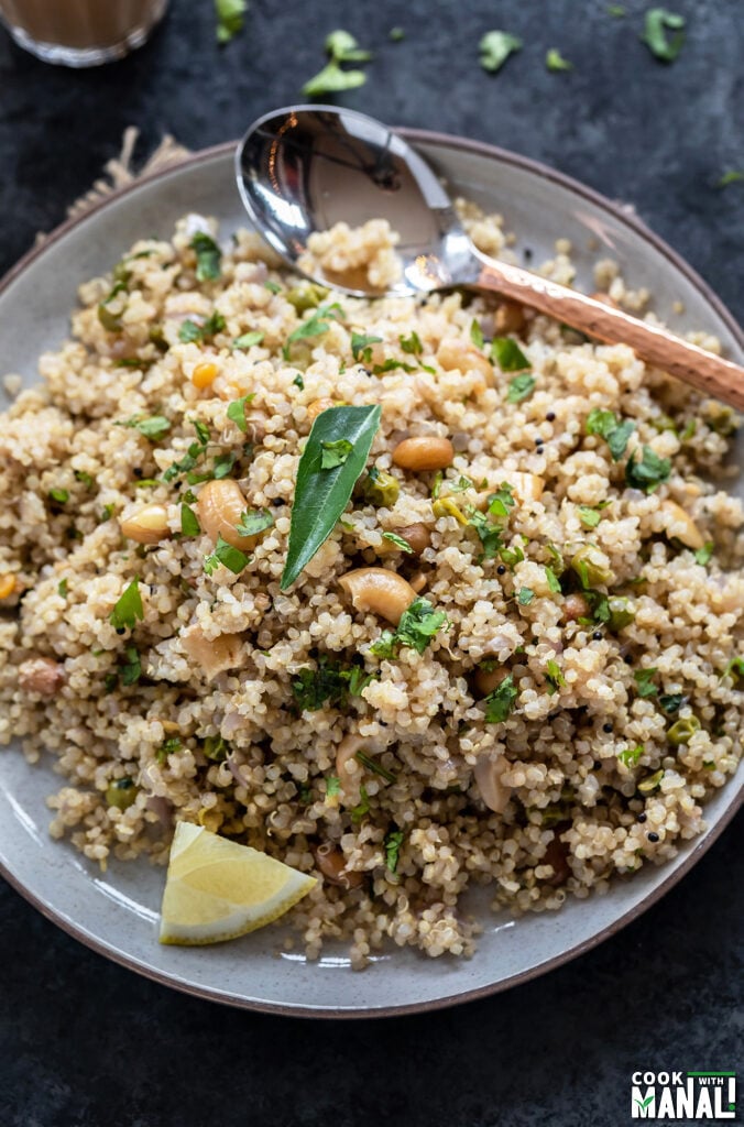 Quinoa upma served in a plate topped with curry leaves, lemon wedge and a copper spoon placed on the side