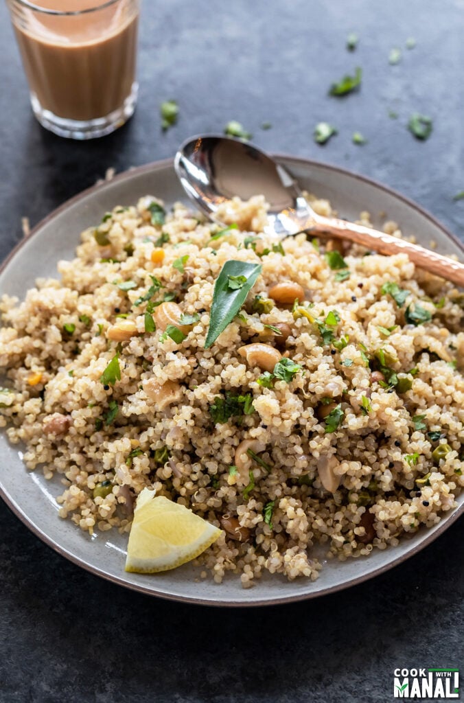 Quinoa upma served in a plate topped with curry leaves, lemon wedge and a copper spoon placed on the side and a glass of chai in the background