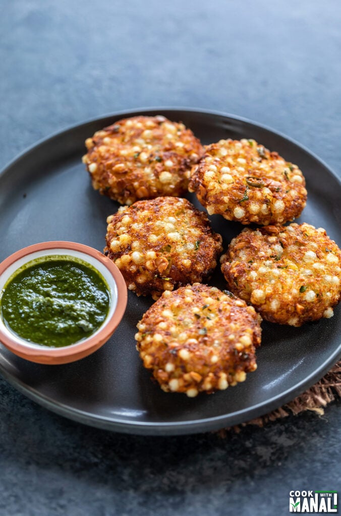 5 pieces of sabudana vada arranged in a plate with a small bowl of cilantro chutney on the side