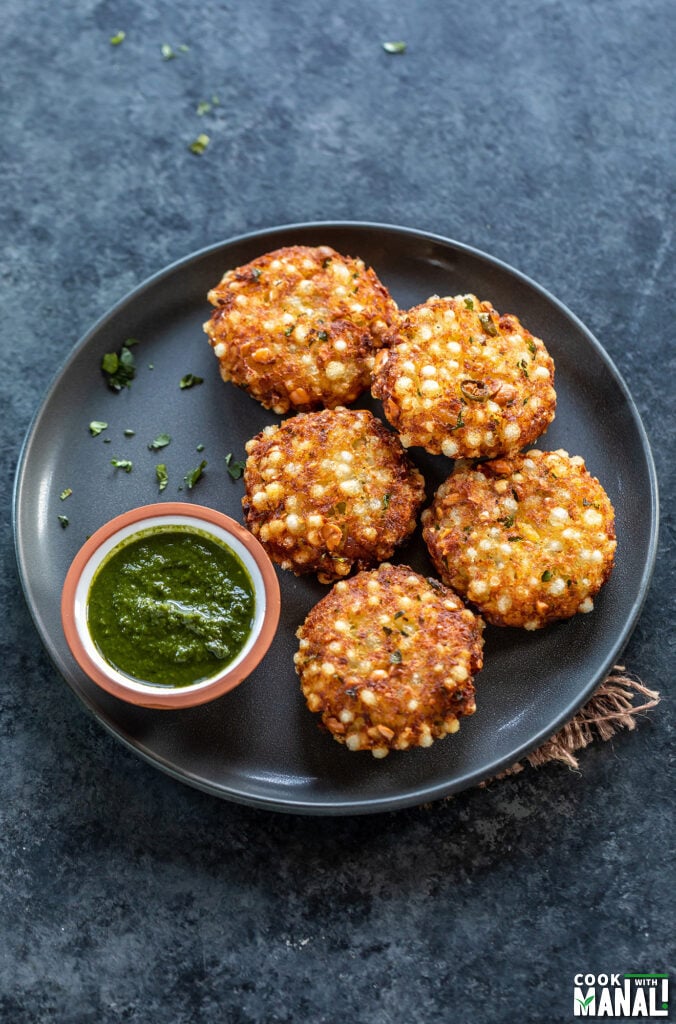 5 pieces of sabudana vada arranged in a plate with a small bowl of cilantro chutney on the side