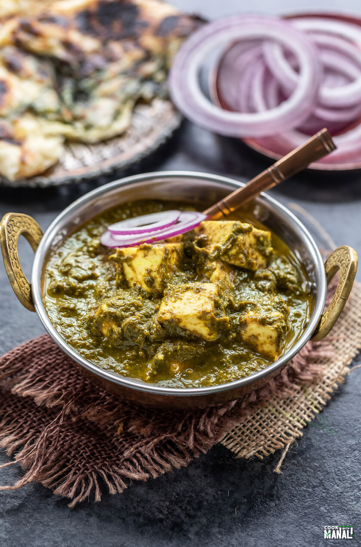 saag paneer served in a small copper kadai with few onion slices placed on one side and a naan placed in the background
