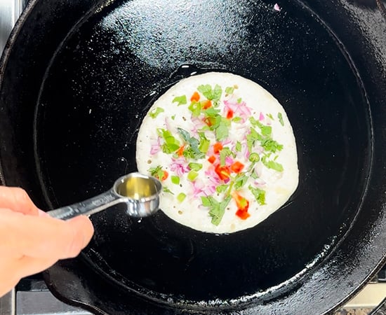 uttapam topped with vegetables and oil being drizzled on the side