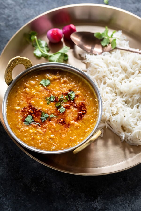 dal served in a copper kadai which is placed in a plate with boiled rice and pickled pearl onions served on the side