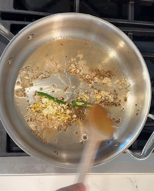 cumin seeds, garlic, ginger being sauteed in a steel pan with a wooden spatula