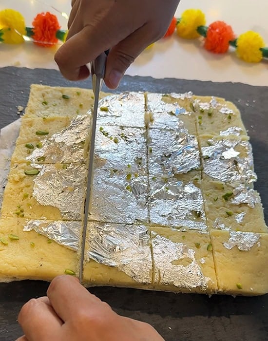 a hand holding a knife cutting burfi into squares