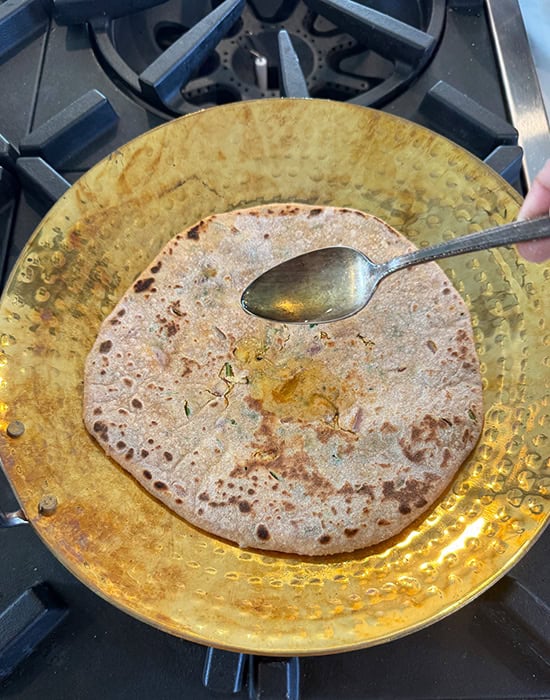 spoon drizzling oil on top of a paratha placed on a tawa