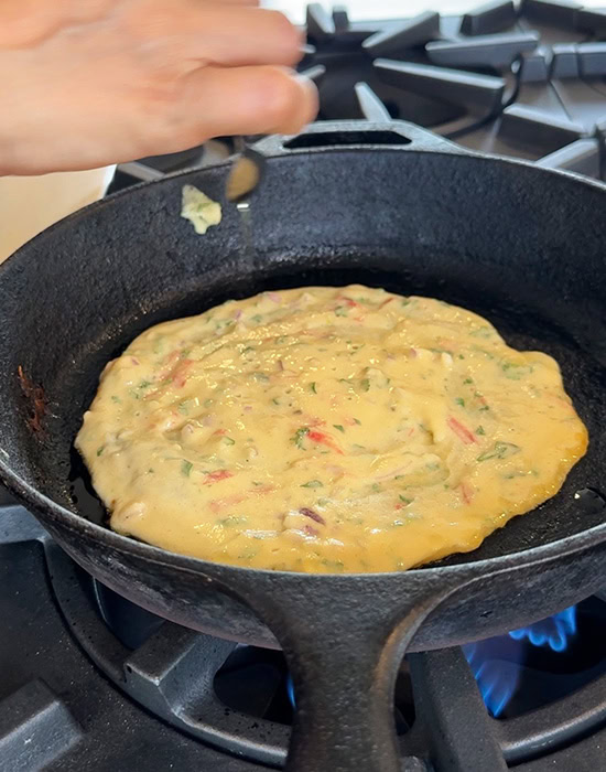 hand adding oil to sides of chilla with a spoon